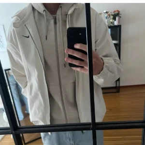 Beige/vit vindjacka från Nike - Säljer en stilren beige vindjacka från Nike med dragkedja och långärmad design. Perfekt för vår och höst. Jackan har en liten Nike-logga på bröstet och en avslappnad passform.