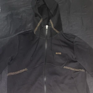 Svart huvtröja från Boss - Snygg svart huvtröja från Boss med dragkedja och justerbar huva. Tröjan har subtila mönsterdetaljer längs ärmarna och runt huvan. Perfekt för en stilren och avslappnad look. Knappt använd. Pris kan diskuteras. står ej för frakt, kan mötas upp i GBG