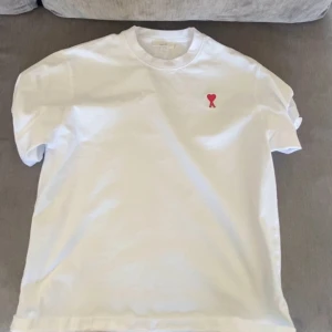 Vit t-shirt med rött hjärta - Säljer en stilren vit t-shirt med ett litet rött hjärta broderat på bröstet. Perfekt för en enkel och snygg look. T-shirten har korta ärmar och en klassisk passform.