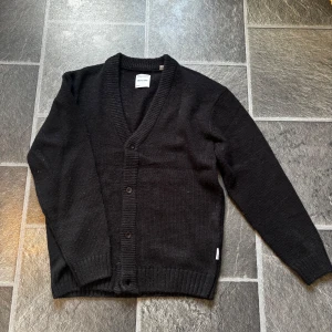 Svart kofta från Only & Sons - Säljer en stilren svart kofta från Only & Sons. Den har en v-ringad design med knappar framtill och är tillverkad i ett mjukt material. Perfekt för att hålla sig varm under kyligare dagar. Sitter perfekt runt 180-185cm! 