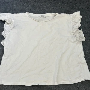 Vit blus från Zara - Säljer en söt vit blus från Zara med volangdetaljer på ärmarna. Perfekt för en stilren och feminin look. Blusen har en rund halsringning och korta ärmar med volanger som ger en extra touch.