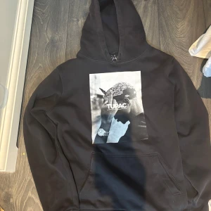 Svart hoodie med Tupac-tryck - Snygg svart hoodie med ett stort tryck av Tupac på framsidan. Perfekt för dig som gillar streetwear och vill ha en bekväm och stilren tröja. Hoodien har en klassisk passform med en stor ficka framtill.