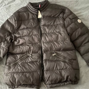 Svart pufferjacka från Moncler - Säljer en stilren svart pufferjacka från Moncler. Jackan har en dragkedja framtill och två snedställda fickor med dragkedjor. Den har en broderad Moncler-logga på ärmen och en ribbad krage. Perfekt för kyligare dagar!