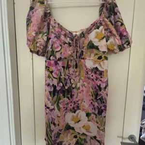 Blommig kortklänning med puffärm XL - Superfin kortklänning med blommigt mönster i rosa, lila, grön och gul. Klänningen har puffärmar, v-ringning med snörning och en luftig passform. Perfekt för sommarens alla tillfällen och ger en romantisk vibe.