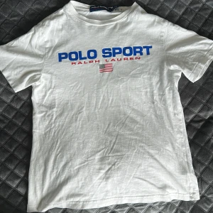 Vit Polo Sport t-shirt från Ralph Lauren - Vit t-shirt från Polo Sport Ralph Lauren med blå och röd logga framtill samt amerikansk flagga. Klassisk rund hals och korta ärmar. Tillverkad i mjuk bomull, perfekt för en clean och sportig look.