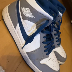 Nike Air Jordan 1 High grå/blå - Snygga Nike Air Jordan 1 High sneakers i grått, vitt och blått. Klassisk high-top siluett med blå snörning och perforerad tå. Ikonisk Air Jordan-logga på sidan och mjuk vadderad plös för extra komfort. Perfekta för dig som gillar streetstyle.