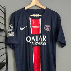PSG matchtröja D. Doué #14 Nike - Snygg PSG fotbollströja i mörkblått med röd och vit rand på framsidan, tryckta detaljer och Qatar Airways sponsor. Officiellt klubbmärke, Champions League-märke på ärmen och D. Doué #14 på ryggen. Tillverkad i lätt och ventilerande material. Helt ny med tagg en replika 