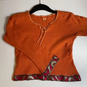 Stickad tröja  - Orange stickad tröja med  dekorativ snörning vid halsen och blommigt band i längs nederkanterna. Tröjan har lätt utsvängda ärmar och är gjord av 100% bomull. Tröjan är köpt secondhand och det står tyvärr ingen storlek men passar S/M. Meddela för mått eller andra frågor, priset kan diskuteras☺️