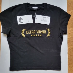 Svart t-shirt Eytys Extra Virgin - Svart t-shirt från Eytys med gul broderad text 'EXTRA VIRGIN' och fem stjärnor framtill. Klassisk rund halsringning och korta ärmar. T-shirten är i bomull och har en clean, rak passform. Perfekt statement-plagg för dig som gillar streetwear.