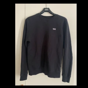 Svart sweatshirt från Vans - En svart sweatshirt från Vans med liten vit logga på bröstet. Klassisk rund halsringning och långa ärmar. Tröjan är i mjuk bomullsmix och har ribbade muddar vid ärmslut och nederkant. 