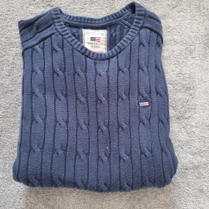 Mörkblå stickad tröja Lexington L - Kabelstickad mörkblå tröja från Lexington i storlek Large. Tröjan har rund halsringning, ribbade muddar och ett litet Lexington-märke på bröstet. Perfekt för dig som gillar klassisk och stilren look med amerikansk preppy-vibe.