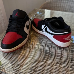 Nike Air Jordan 1 Low svart/röd/vit - Säljer ett par Nike Air Jordan 1 Low i klassisk svart, röd och vit färg. Skorna har läderöverdel, svart swoosh och röda detaljer på tå och häl. Ikonisk Jordan-logga på plösen och vinge på hälen. Nypris 1600kr, använt 3 gånger. Tvättar dom rent + nya snören skickas vid säljning. 