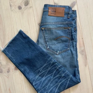 G-Star RAW 3301 blå jeans straight fit - Snygga blå jeans från G-Star RAW, modell 3301, med klassisk femficksdesign och raka ben. Jeansen har slitna detaljer, synliga knappar i gylfen och råa, avklippta benslut vilket gör de lite kortare. Det är originellt 32 i längd men skulle nu säga att det är 29-30. Galet snygg wash och bra kvalitet!!
