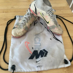 Nike Mercurial fotbollsskor vit/rosa - Säljer ett par Nike Mercurial fotbollsskor i vit och ljusgrå med rosa och svarta detaljer. Skorna har snörning, platt sula med dobbar och är tillverkade i syntetmaterial med flyknit-överdel. Swoosh-logga på sidan och cool design för dig som vill sticka ut på planen.