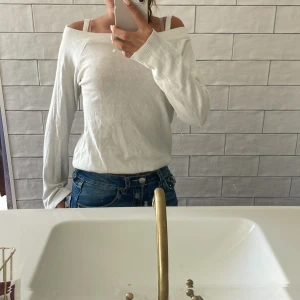 Vit offshoulder långärmad tröja - Vit långärmad tröja med offshoulder-modell och bred ribbad kant upptill. Tröjan har smala axelband och är tillverkad i ett mjukt, tunt material som känns skönt mot huden. Perfekt för dig som gillar en stilren och enkel look med en twist.