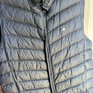 Svart dunväst från Gant  - Säljer en svart dunväst från Gant, modell Airlight Vest. Västen har quiltad design med horisontella sömmar, dragkedja framtill och Gant-logga med sköld på bröstet. Perfekt att slänga över en hoodie eller tröja för extra värme.