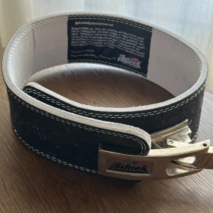 Svart träningsbälte från Schiek - Säljer ett svart träningsbälte från Schiek med kraftigt spänne i metall och vit kontrastsöm. Bältet är brett och tillverkat i slitstarkt skinn, perfekt för dig som vill ha extra stöd vid tunga lyft. Insidan är ljus och har en tydlig varningsetikett.