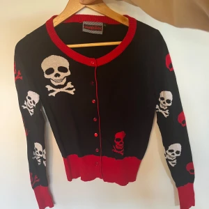 Svart kofta med dödskallemönster - Svart kofta med röda och vita dödskallar över hela tröjan. Röd ribbad kant vid hals, knäppning och nederkant. Långärmad och figurnära passform, knäpps med röda knappar framtill. Asnajs🔥🔥☠️☠️ Unik därav priset❤️aldrig använd!