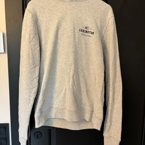 Grå Lexington sweatshirt i bomull M - Snygg grå sweatshirt från Lexington i 100% bomull. Tröjan har broderad Lexington-logga med flagga på bröstet och klassisk rund hals. Perfekt basic med lång ärm och ribbade muddar för en clean och avslappnad look.