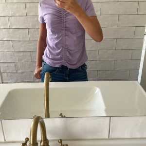 Ljuslila topp med rynk från ONLY - Superfin ljuslila topp från ONLY med korta ärmar och v-ringning. Toppen har snygg rynkdetalj längs hela framsidan som ger en cool och trendig look. Perfekt till jeans eller kjol för en chill stil. Mjuk och skön i bomull.