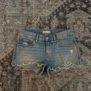 Blå lågmidjade jeansshorts  - Snygga blå jeansshorts från Perfect Jeans med råa, fransiga kanter och slitna detaljer framtill. Klassisk femficksmodell med knapp och dragkedja. Perfekta för varma dagar när du vill ha en avslappnad och trendig look.