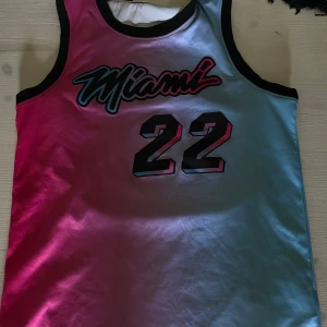 Miami Heat Jimmy Butlrt basketlinne - Snyggt basketlinne med Miami Heat och Jimmy Butler tryck. Linnet har en cool färgövergång från rosa till ljusblått och svarta detaljer runt ärmhål och hals. Perfekt för dig som gillar basket och streetstyle.