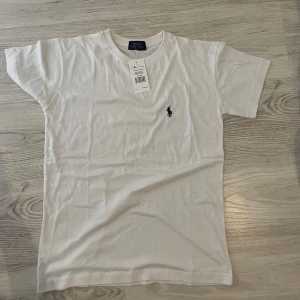 Vit t-shirt från Polo Ralph Lauren - Vit t-shirt från Polo Ralph Lauren med klassisk passform och korta ärmar. Tillverkad i mjuk bomull och pryds av den ikoniska broderade loggan i mörkblått på bröstet. Enkel och stilren design som funkar till allt.