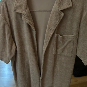 Beige kortärmad frottéskjorta - Mjuk och chill beige skjorta i frotté med korta ärmar, klassisk krage och en bröstficka. Perfekt för en avslappnad look och enkel att styla med jeans eller shorts.