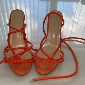 Orange sandaler med snörning och klack - Trendiga orange sandaler med blockklack och snygg snörning som går att vira runt benet. Skorna har öppen tå och en cool, präglad struktur som påminner om krokodil. Perfekta för att lyfta din sommarlook.