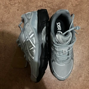Asics GEL NYC - Snygga Asics GEL sneakers i grått och silver med svarta detaljer på sulan. Skorna har mesh och syntetpaneler, klassisk snörning och rund tå. Perfekta för dig som gillar sportig och trendig stil med retrovibbar.
