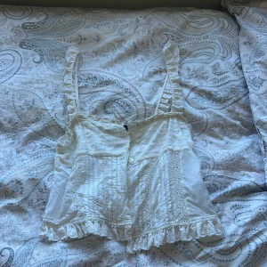 Vit volangblus från Zara XS - Supersöt vit blus från Zara i storlek XS med volangdetaljer på axelband och nederkant. Blusen har knappar framtill, spetsinslag och romantiska detaljer. Perfekt för dig som gillar en feminin och vintageinspirerad look.