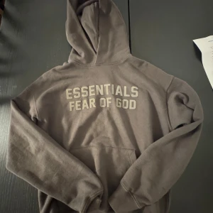 Essentials Fear of God tracksuit  - Säljer en brun hoodie från Essentials Fear of God med stor logga på bröstet. Tröjan har huva, känguruficka och långa ärmar. Perfekt för dig som gillar streetwear och vill ha en clean look med snygg branding.