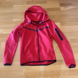 Röd Nike Tech  - Röd vindjacka från Nike med svarta detaljer och hel dragkedja. Jackan har huva, lång ärm och en praktisk ficka med dragkedja på ena ärmen. Perfekt för dig som gillar sportig stil och vill ha något lätt och snyggt till vardags.