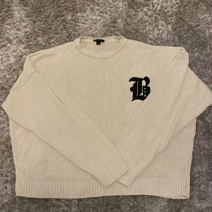 Beige stickad tröja med svart B-logga - Beige stickad tröja från Boohoman med en stor svart B-logga på bröstet. Tröjan har rund halsringning, ribbade muddar och är långärmad. 