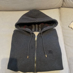 Mörkgrå Burberry hoodie med dragkedja - Snygg mörkgrå hoodie från Burberry med klassiskt rutigt foder i huvan och guldfärgade detaljer på snörena. Tröjan har dragkedja framtill, fickor och broderad logga på bröstet. Tillverkad i mjuk bomull och polyester för extra komfort.