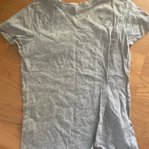 Grå t-shirt från Hollister - En basic grå t-shirt från Hollister i 100% bomull. Klassisk passform med rund hals och korta ärmar. Diskret Hollister-logga på bröstet. Perfekt till jeans eller shorts för en chill look.