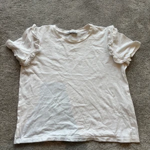 Vit volangtopp från Zara - Säljer en söt vit topp från Zara med korta ärmar och volangdetaljer på axlarna. Toppen har en klassisk rund halsringning och är gjord i mjuk bomull, perfekt för en chill och stilren look.