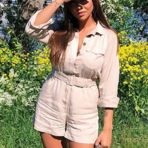 Beige playsuit med bälte och knappar - Snygg beige playsuit med långa ärmar, knappar framtill och tillhörande bälte i midjan. Modellen har bröstfickor och uppvikta ärmslut, perfekt för varma dagar. Lätt och bekväm, passar dig som gillar en stilren look.