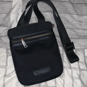 Svart axelväska från Valentino - Snygg svart axelväska från Valentino med justerbar rem och dragkedja i metall. Väskan har ett stilrent utseende med diskret logga framtill och extra innerfack med dragkedja. Perfekt storlek för mobil, plånbok och andra essentials.