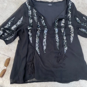 Svart blus med paljetter från KappAhl - Svart blus från KappAhl med glittriga paljettmönster i silver längs ärmar och framtill/baktill. Blusen har v-ringning med knytband och tofsar. Perfekt för dig som vill sticka ut med en cool och unik look.