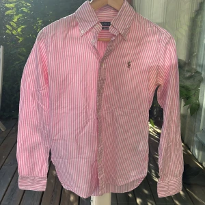 Rosa skjorta Ralph Lauren - Säljer en klassisk rosa- och vitrandig skjorta från Ralph Lauren med broderad logga på bröstet. Snygg slim fit-modell i mjuk bomull, perfekt för en clean och fräsch look. Sitter rätt så slim passar xs / s perfekt för sommaren