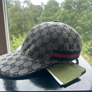 Gucci keps  - Snygg keps från Gucci i beige med klassiskt GG-mönster över hela kepsen. Har en röd och marinblå rand på sidan och justerbar rem baktill. Perfekt accessoar för att lyfta din outfit med lyxig känsla.