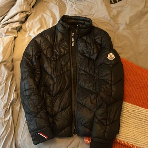 Svart Moncler dunjacka  - Säljer en svart Moncler dunjacka med klassisk logga på ärmen och snygg quiltad design. Jackan har hög krage, dragkedja framtill och coolt serietryck på insidan. Perfekt för kalla dagar och riktigt stilren look.