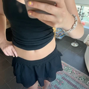 Svart volangkjol - Supersöt svart kortkjol med smock i midjan och dubbla volanger. Kjolen är luftig och har en lätt transparent känsla i nederdelen. Perfekt för dig som gillar en enkel men ändå trendig look.