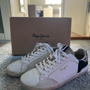 Pepe Jeans vita sneakers strl 41 - Snygga vita sneakers från Pepe Jeans med svarta och beige detaljer. Klassisk låg modell med rund tå, snörning och diskret logga i guld på sidan. Ovandel i skinn och textil, perfekt för en clean och stilren look.