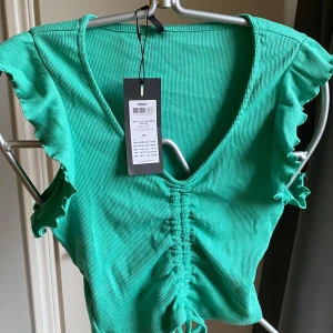 Grön ribbad topp med volang från ONLY! Ny - ! Supersöt grön ribbad topp från ONLY med volangdetaljer på ärmarna och snygg snörning framtill, den är justerbar! Toppen har v-ringning och är croppad, perfekt för en trendig look. Materialet är mjukt och stretchigt, vilket gör den riktigt bekväm att bära. 