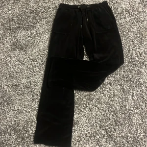Svarta velourbyxor Juicy Couture - Svarta mjukisbyxor från Juicy Couture i mjuk velour med broderad JC-logga framtill. Byxorna har dragsko i midjan, raka ben och fickor både fram och bak med knappdetaljer. Perfekta för en chill och trendig look. Andvända endast fåtal gånger. Ordenarie pris 1100