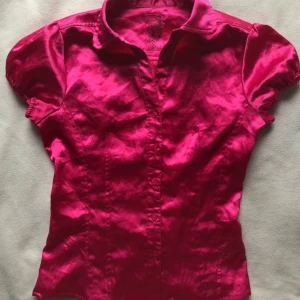 Glansig rosa skjorta från Windy Girls - Snygg glansig rosa skjorta från Windy Girls med korta puffärmar, klassisk krage och knappar framtill. Baksidan har cool snörning som ger en unik look. Perfekt för dig som vill sticka ut med färg och detaljer.