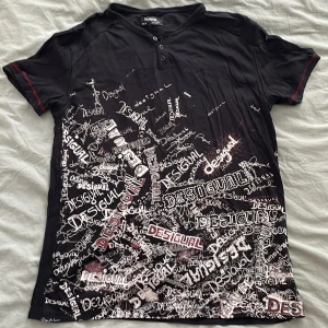 Svart t-shirt med tryck från Desigual - Cool svart t-shirt från Desigual med massor av vita och röda logotyper tryckta över hela framsidan. T-shirten har korta ärmar, knappar vid halsen och röda sömdetaljer på ärmarna. Skön passform och tillverkad i mjuk bomull.