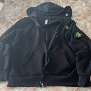 Svart hoodiejacka från Stone Island - Svart hoodiejacka från Stone Island med dragkedja, huva och klassisk logopatch på ärmen. Jackan har två stora fickor framtill och ribbade muddar. Tillverkad i mjuk bomull, perfekt för en chill och stilren look.